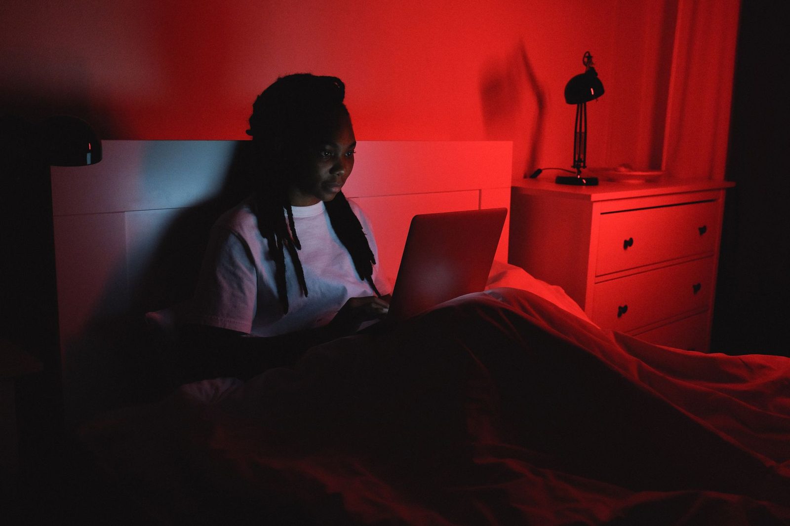 Giovane donna seduta a letto di notte davanti a un computer portatile, in una camera illuminata di rosso
