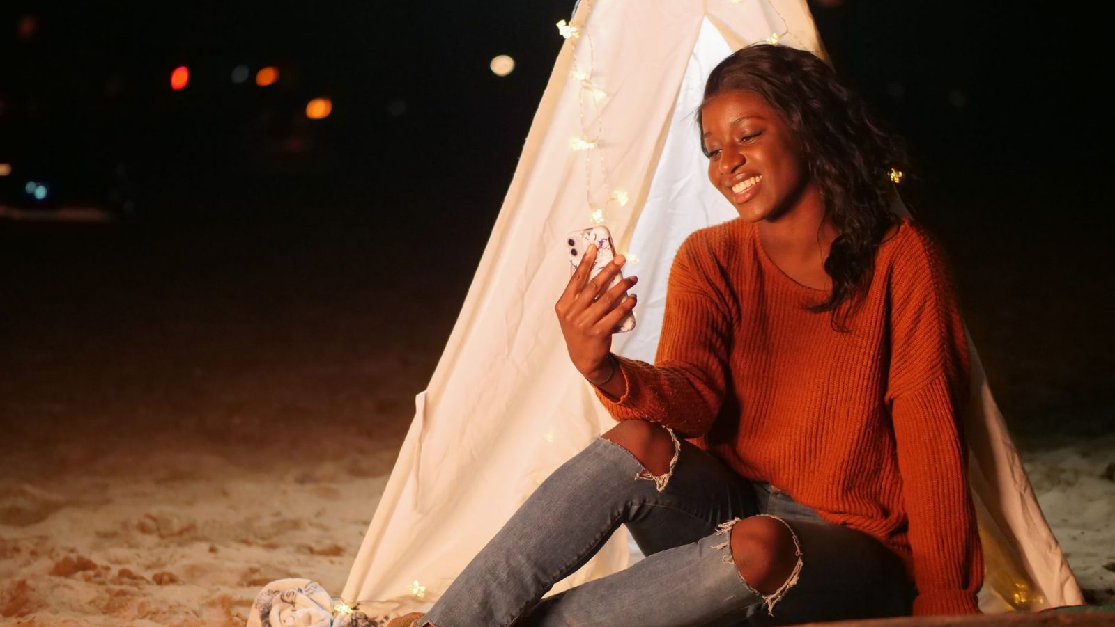 Smiling young woman in an orange sweater looking at her smartphone, sitting outside a glowing tent at night