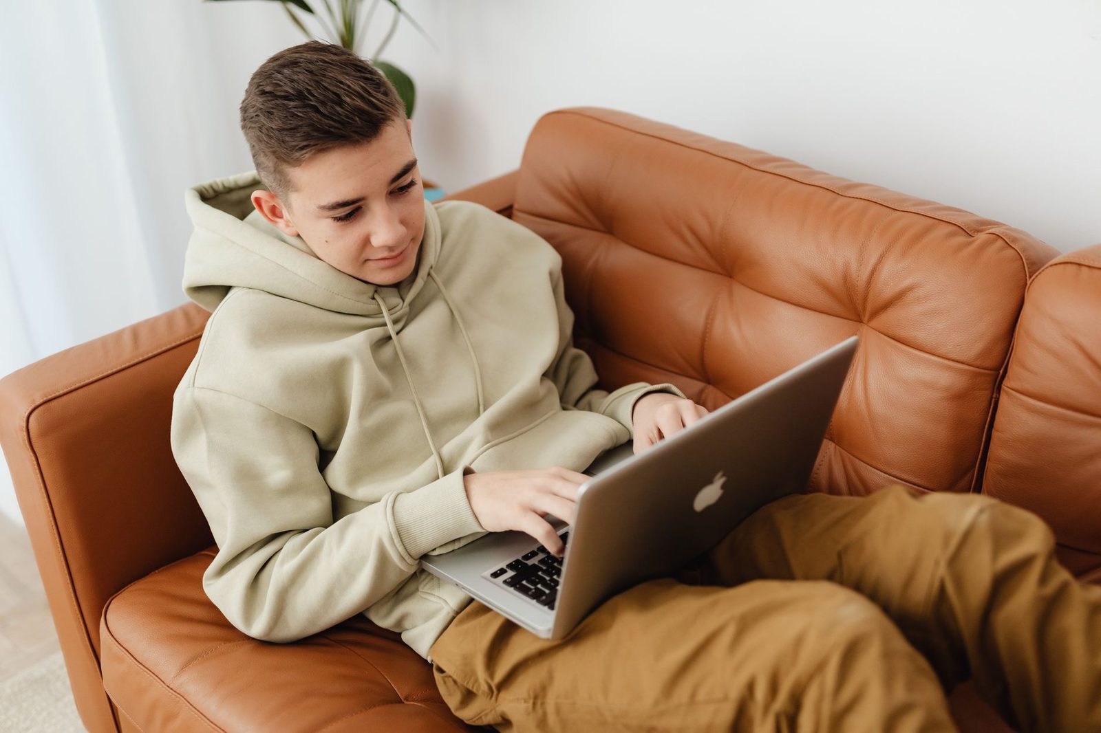 Junger Mann im Kapuzenpullover sitzt auf einem braunen Ledersofa und benutzt einen Laptop