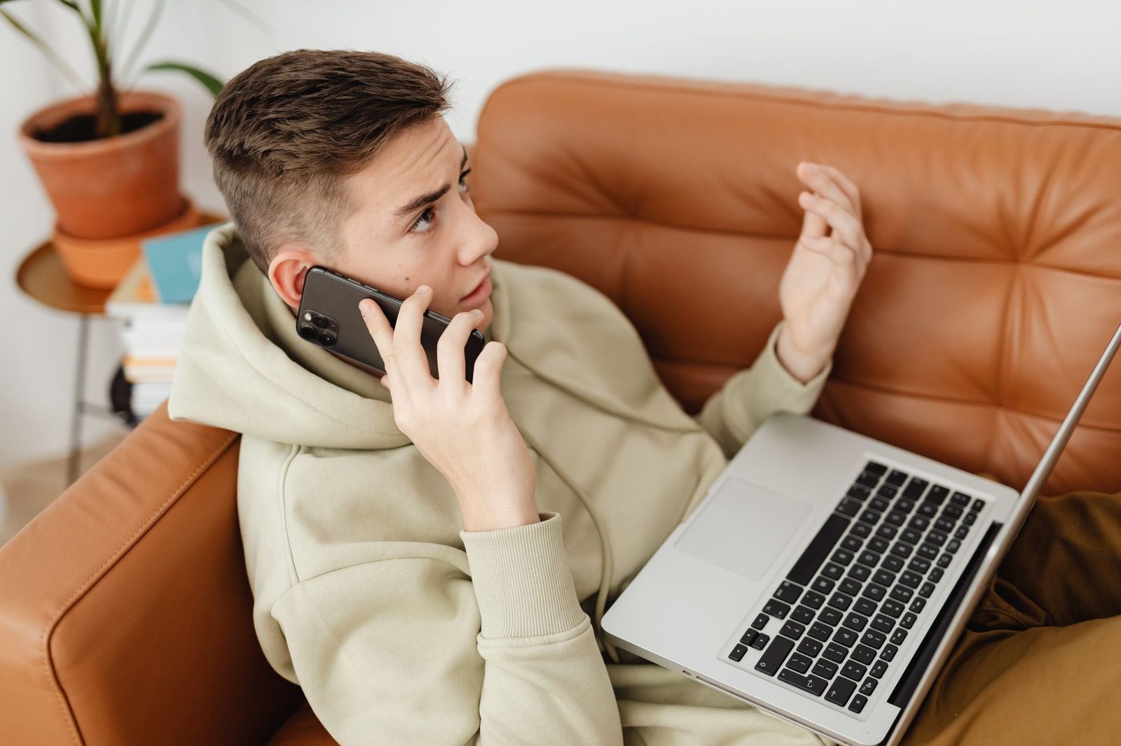 Jeune homme en sweat assis sur un canapé en cuir, téléphone à l'oreille et ordinateur portable sur les genoux