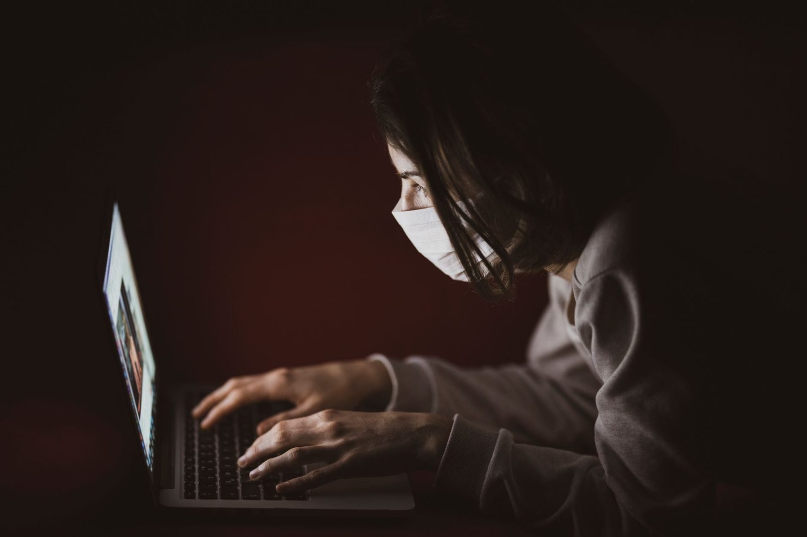 Mujer con mascarilla quirúrgica inclinada sobre un ordenador portátil en una habitación oscura, con la pantalla encendida