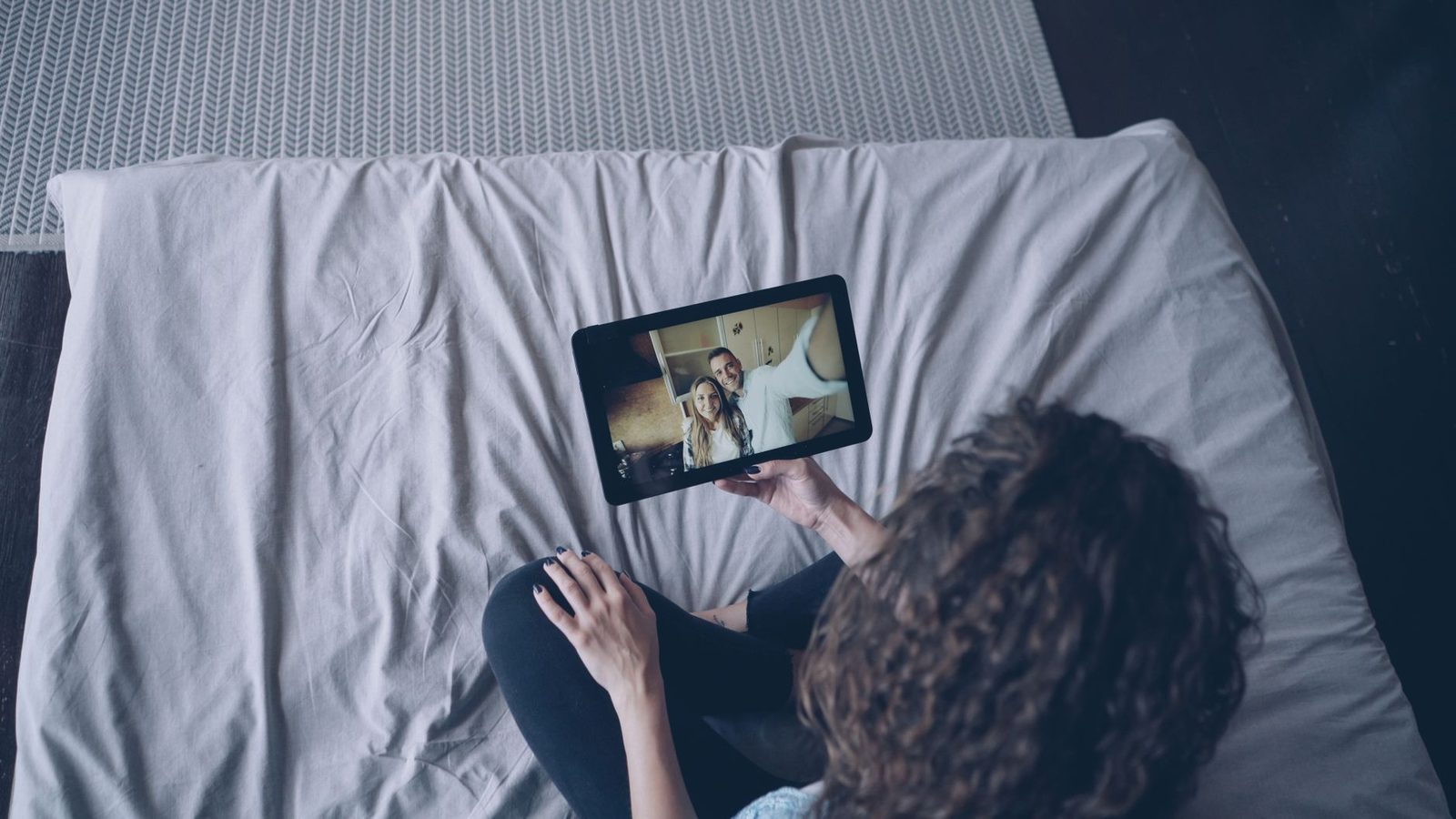 Femeie așezată pe un pat ținând o tabletă în timpul unui apel video cu un cuplu pe ecran