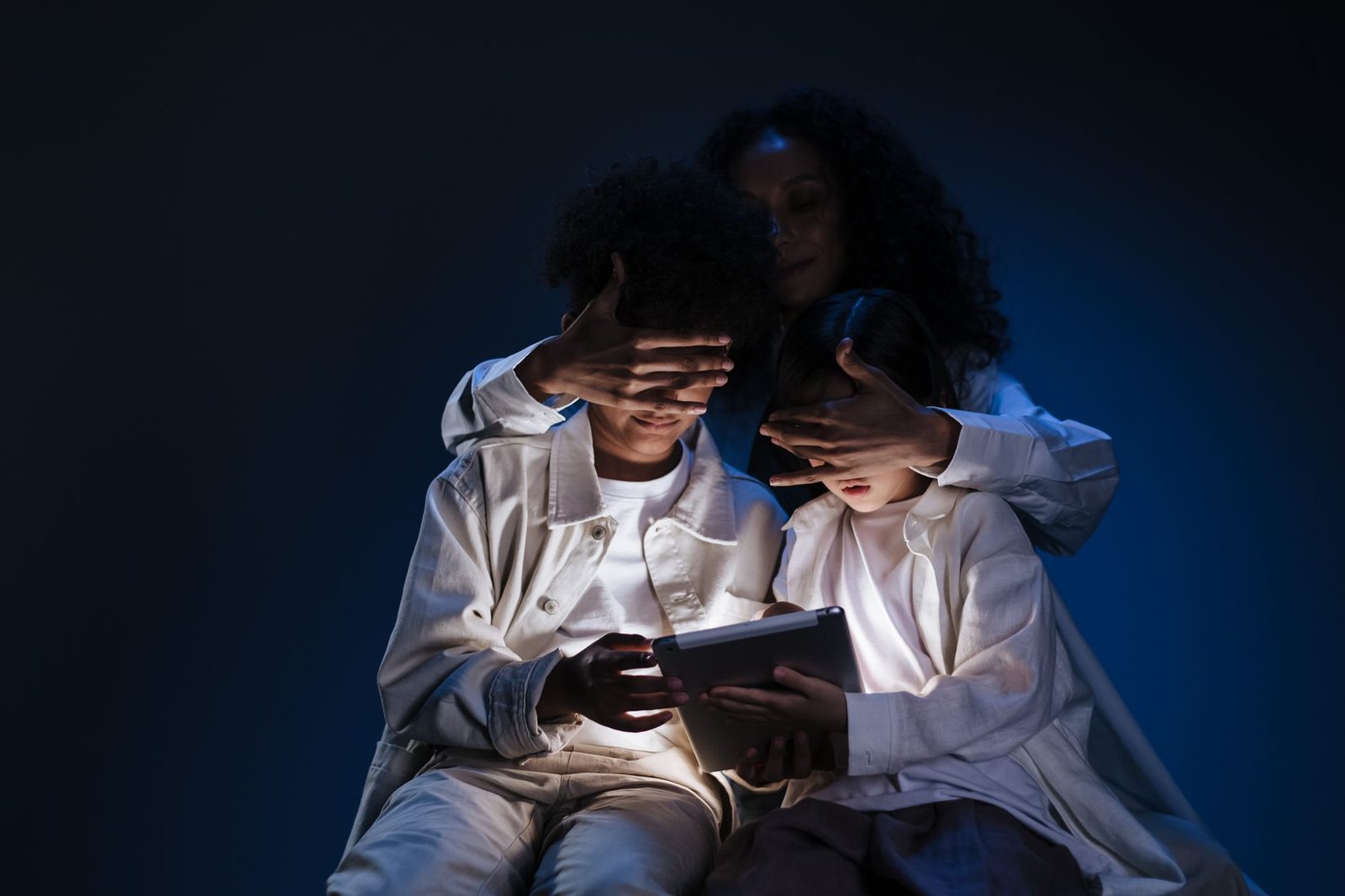 Une femme cache les yeux de deux enfants devant l'écran lumineux d'une tablette dans l'obscurité