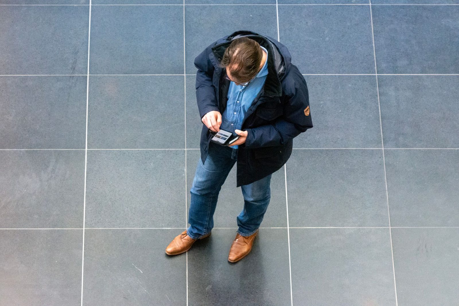 Vue de dessus d'un homme en manteau et jean consultant son smartphone, debout sur un sol carrelé gris