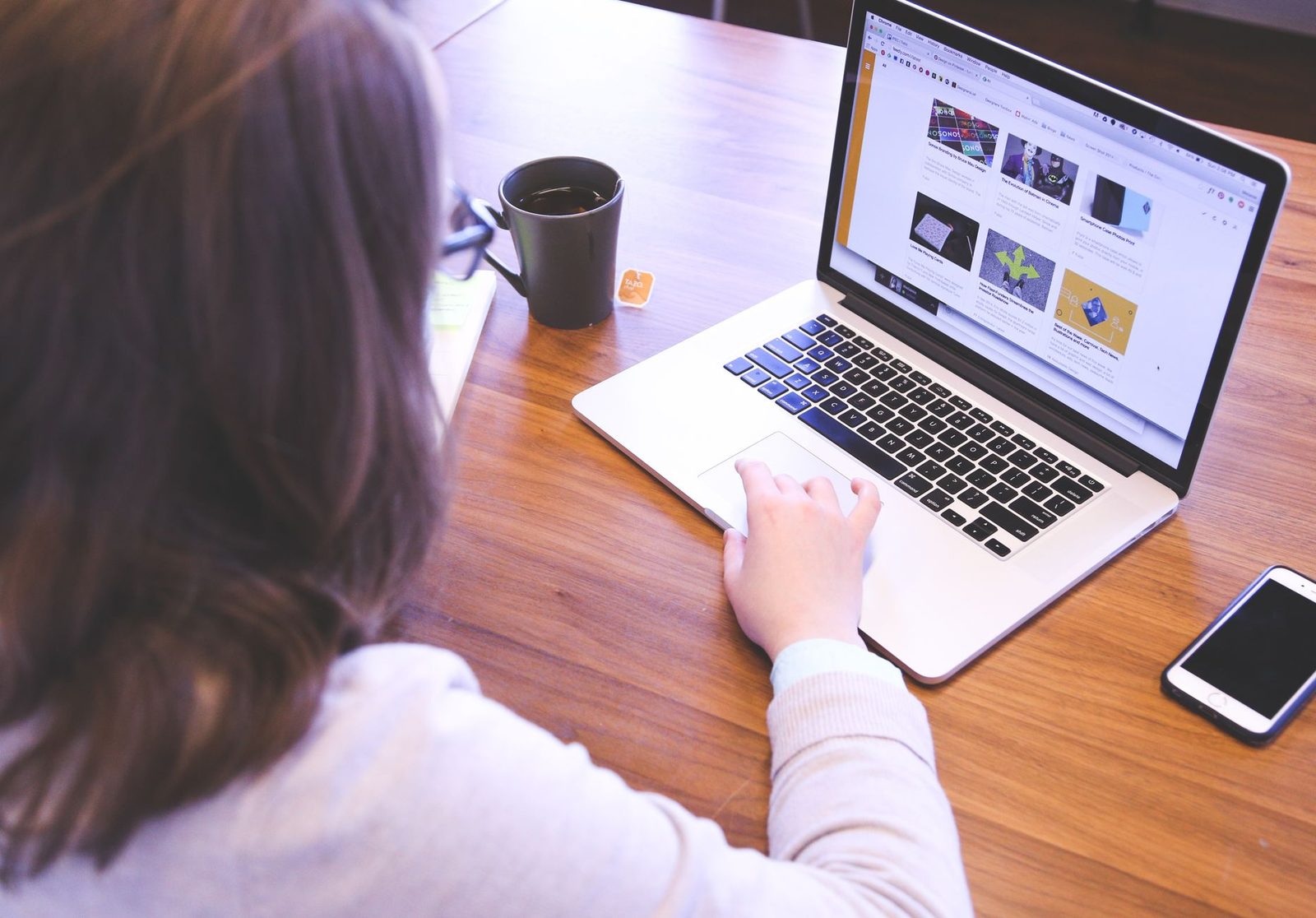 Femme assise à une table en bois naviguant sur un site web depuis un ordinateur portable, tasse et smartphone à côté