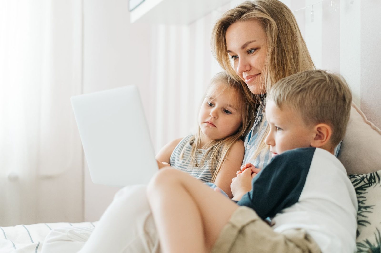 Une mère et ses deux jeunes enfants regardent ensemble l'écran d'un ordinateur portable sur un lit
