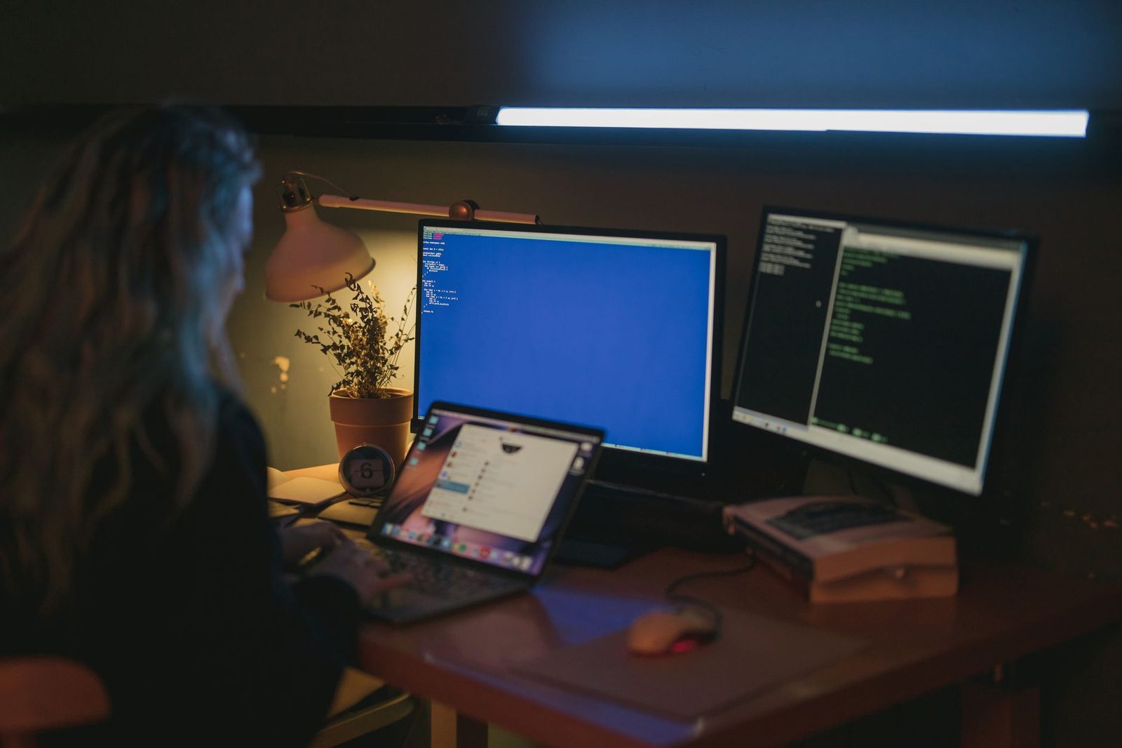 Personne de dos devant un bureau sombre avec plusieurs écrans d'ordinateur allumés et un ordinateur portable