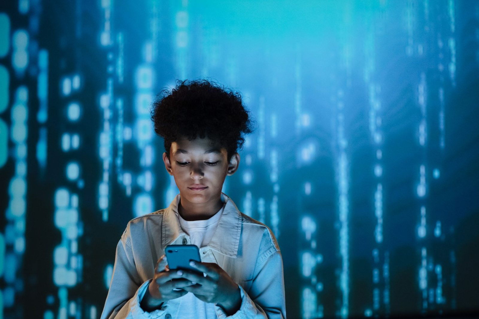 Adolescent consultant un smartphone devant un mur lumineux bleu couvert de données numériques