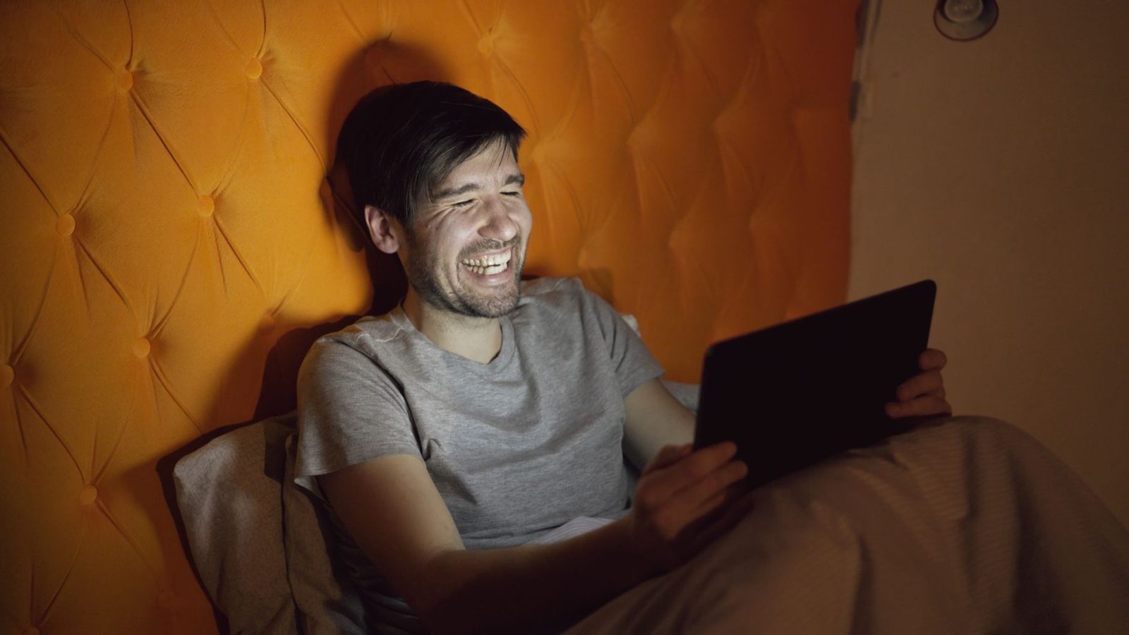 Homme souriant allongé dans un lit, consultant une tablette numérique dans une chambre faiblement éclairée