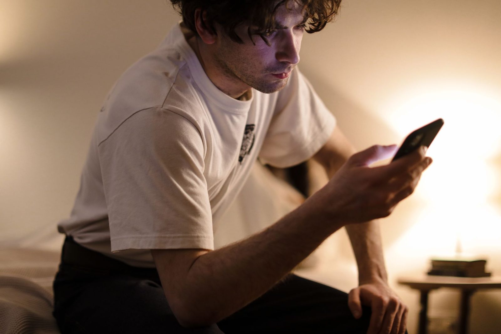 Jeune homme assis sur un lit consultant son smartphone dans une chambre faiblement éclairée le soir