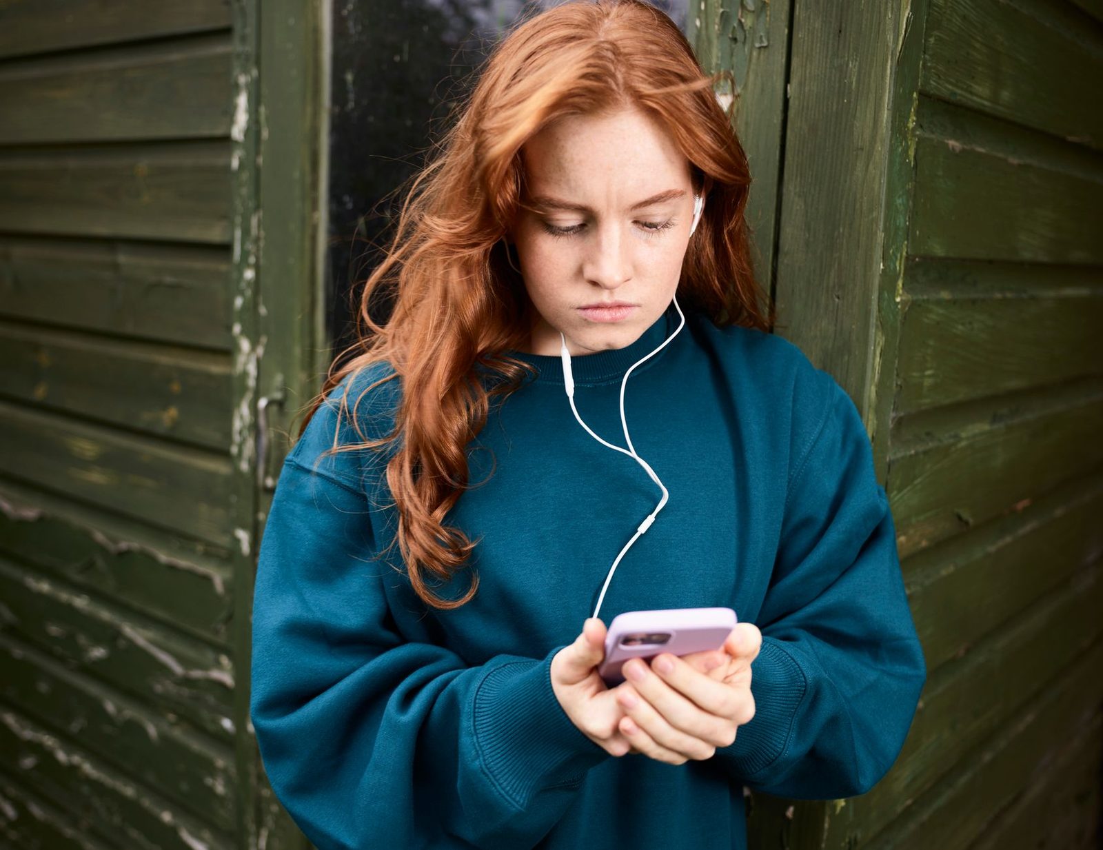 Adolescente rousse avec écouteurs filaires regardant son smartphone devant une porte en bois verte